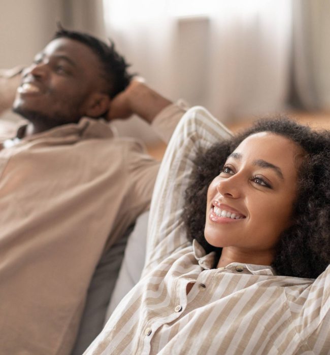 Weekend bliss. Relaxed black millennial couple leans back on their living room sofa indoor, posing with smiles and holding hands above their heads. Comfort and love. Selective focus